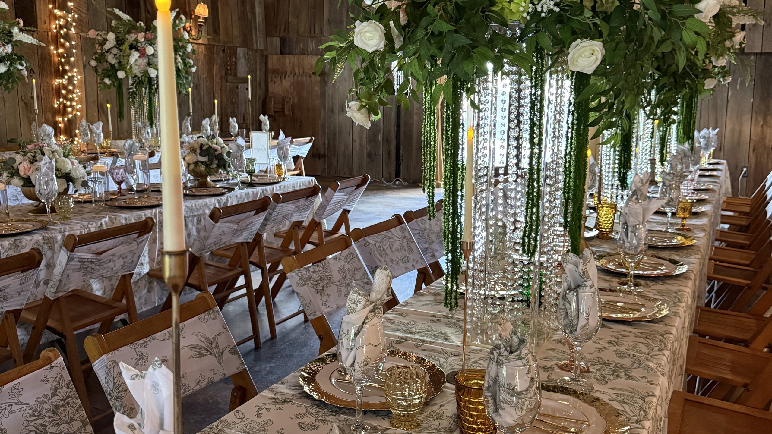 Long dinner tables set inside the barn with gold chargers, candles and greenery, string lights on the barn walls