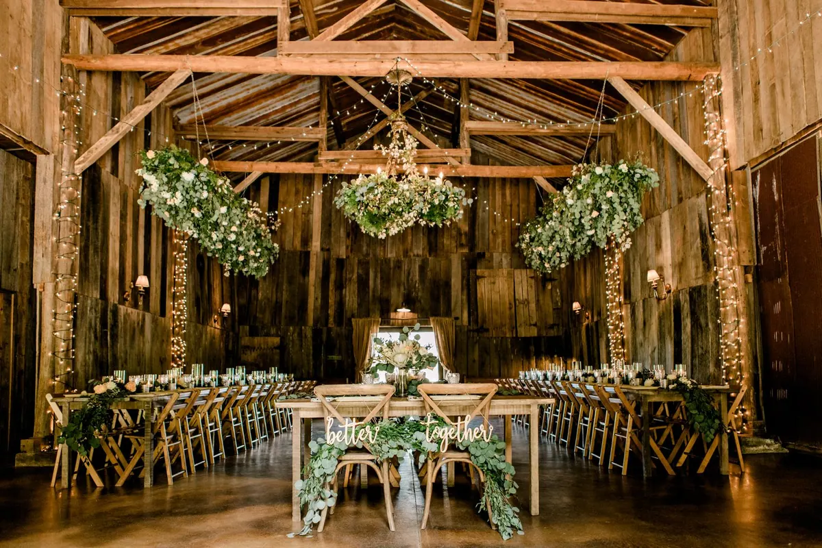Long farm tables set for dinner inside the barn, greenery and chandeliers overhead